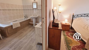 Un baño con bañera, lavamanos y espejo. Hay una mesita de noche de madera con lámpara y una cama con colcha estampada.