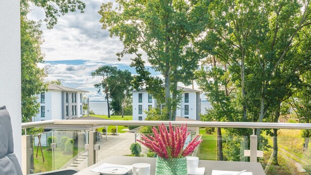 A dining table with a bowl of fruit, white cups, and a green vase with pink flowers, overlooking a pool and buildings. - Apartment sun deck - Haffresidenz (4a.12) - 4a.12 (Zirchow)