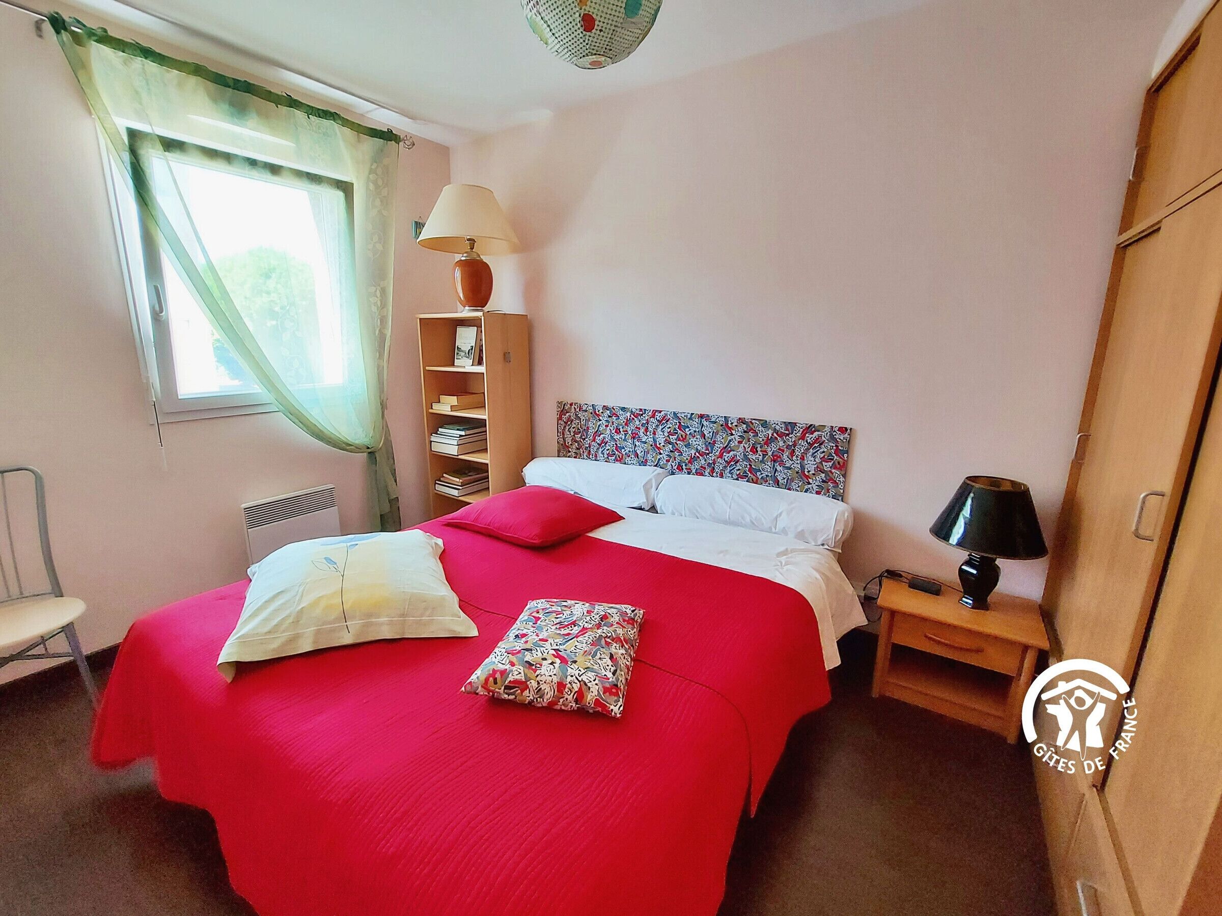A bedroom with a bed, a nightstand, a lamp, a bookshelf, a chair, and a window with curtains.