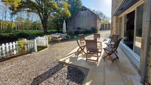 An outdoor patio with a table and chairs, a white picket fence, and a rustic barn in the background. - Riverside Retreat: Spacious 3BR House, Private Fishing Pond, Near Charming Villages & Outdoor Activities (Voutré)