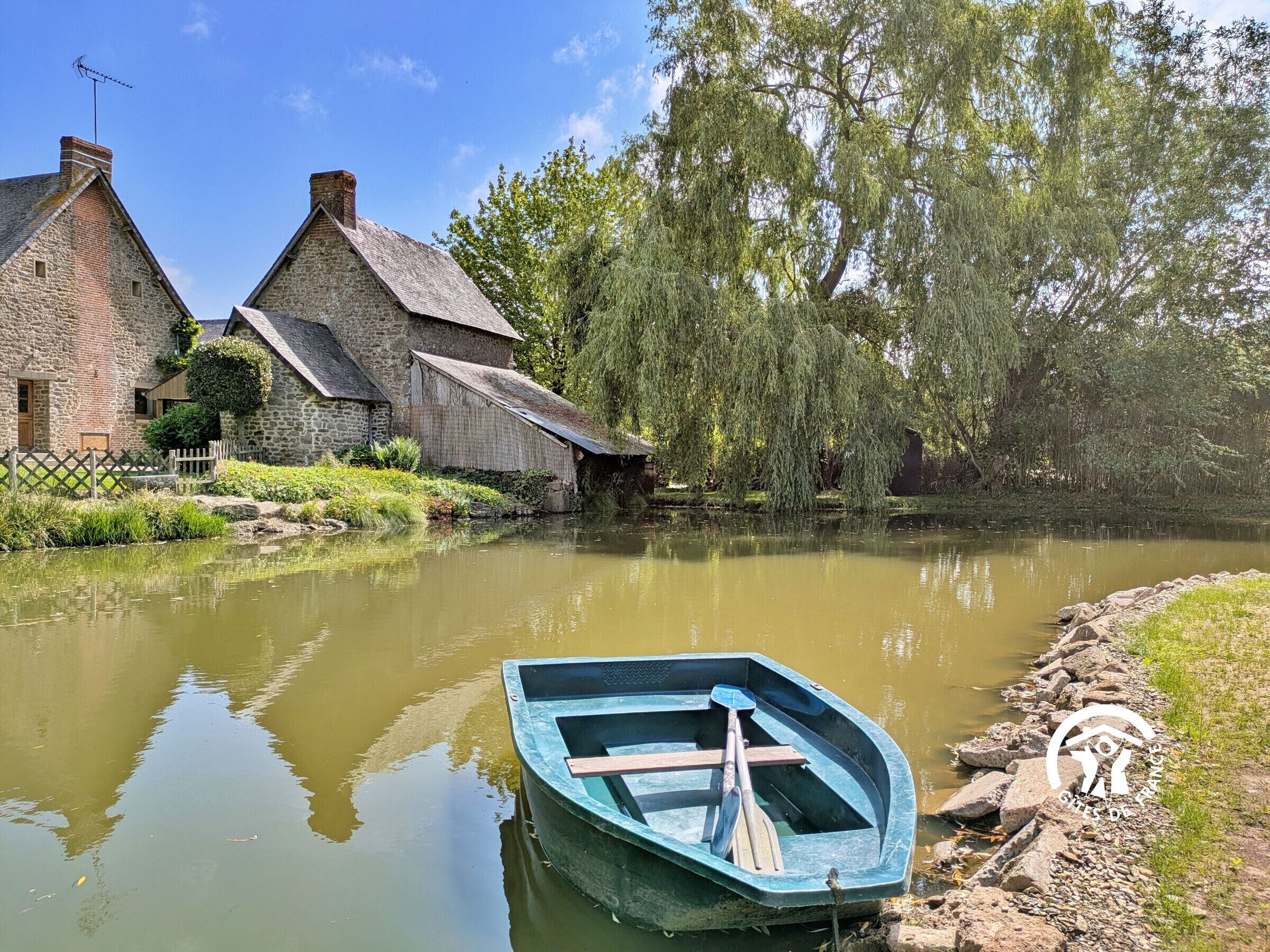 A rustic stone building with a thatched roof, a small boat on the water, and a weeping willow tree.