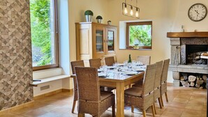 A dining room with a wooden table set for six, surrounded by wicker chairs. There is a window with a view of greenery, a cabinet with glass doors, and a fireplace with a clock above it.