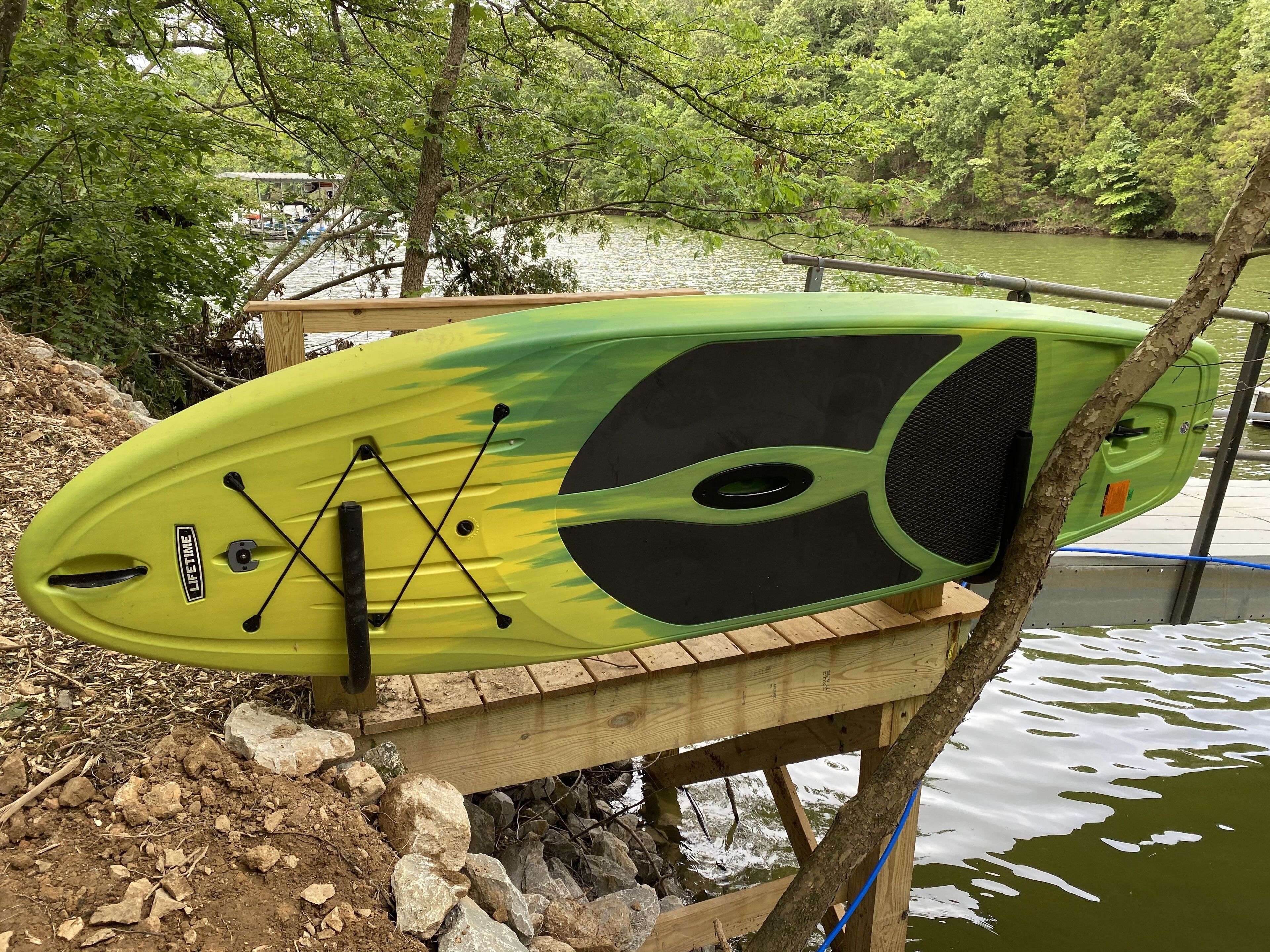 Un kayak con diseño negro y verde, sujeto a un muelle de madera cerca de un árbol.