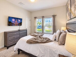 Quarto com cama, cômoda, televisão na parede e janela com cortinas.