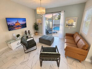 A modern living room with a flat-screen TV, a glass coffee table, a leather sofa, and a dining area with a white cabinet.