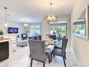Uma sala de jantar moderna com uma mesa grande, cadeiras e um candelabro. Há uma televisão na parede e uma janela com vista para o verde.