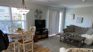 A living room with a dining table, chairs, a sofa, a coffee table, a television, and a large window with blinds. - 2 BR, 1 bath w full kitchen on ground level w full kitchen w access to amenities (Murrells Inlet)
