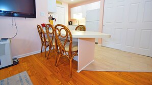 A compact kitchen with white cabinets, a built-in refrigerator, a dining area with wooden chairs, and a flat-screen TV mounted on the wall.