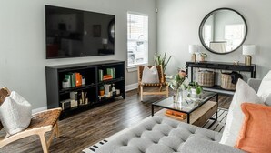 A modern living room with a flat-screen TV, a black entertainment unit, a round mirror, and a comfortable seating area with a grey sofa and a wooden chair. - Brand New and One Block from Magnolia Silos! (Waco)
