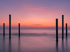 Une étendue d’eau calme, parsemée de piliers en bois qui s’y avancent, sous un ciel embrasé par le coucher du soleil.