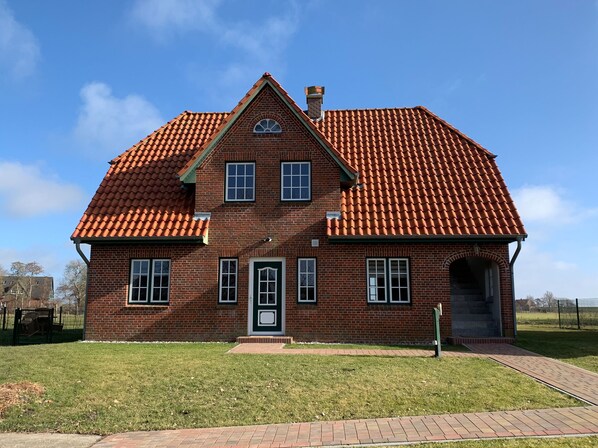 Une maison à deux étages, construite en briques, avec un toit recouvert de tuiles rouges, une cheminée centrale et une porte d’entrée dotée d’un cadre vert.