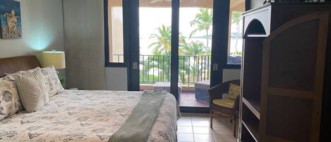 A bedroom with a bed, a nightstand, a TV cabinet, and a balcony with a view of palm trees.