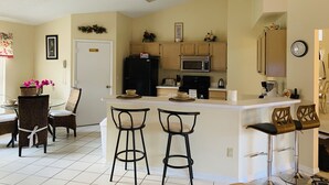 Una cocina con isla central, taburetes de bar, zona de comedor con sillas de mimbre y un refrigerador.