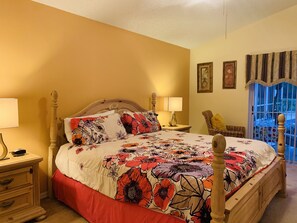 A bedroom with a floral bedspread, wooden headboard, bedside lamps, a wicker chair, and a window with a view.