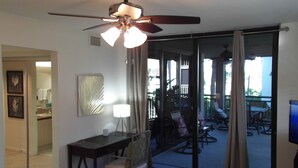 Une chambre d’hôtel équipée d’un ventilateur de plafond, d’un bureau avec une chaise, d’une grande fenêtre avec des rideaux et donnant sur l’espace extérieur aménagé avec des sièges.