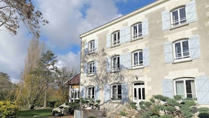 Un edificio de varios pisos con fachada simétrica, amplias ventanas con contraventanas y un jardín bien cuidado con arbustos diversos y césped.