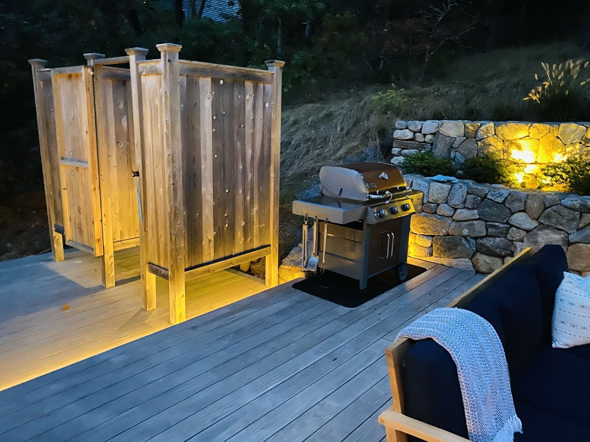 Een terras buiten met houten vloerdelen, een barbecue en een stenen muur met verlichting.