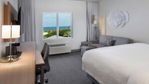 Habitación de hotel con cama, escritorio, silla, televisor, ventana con vista al océano y un adorno en la pared.