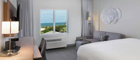 A hotel room with a bed, a desk, a chair, a TV, a window with a view of the ocean, and a decorative wall piece.