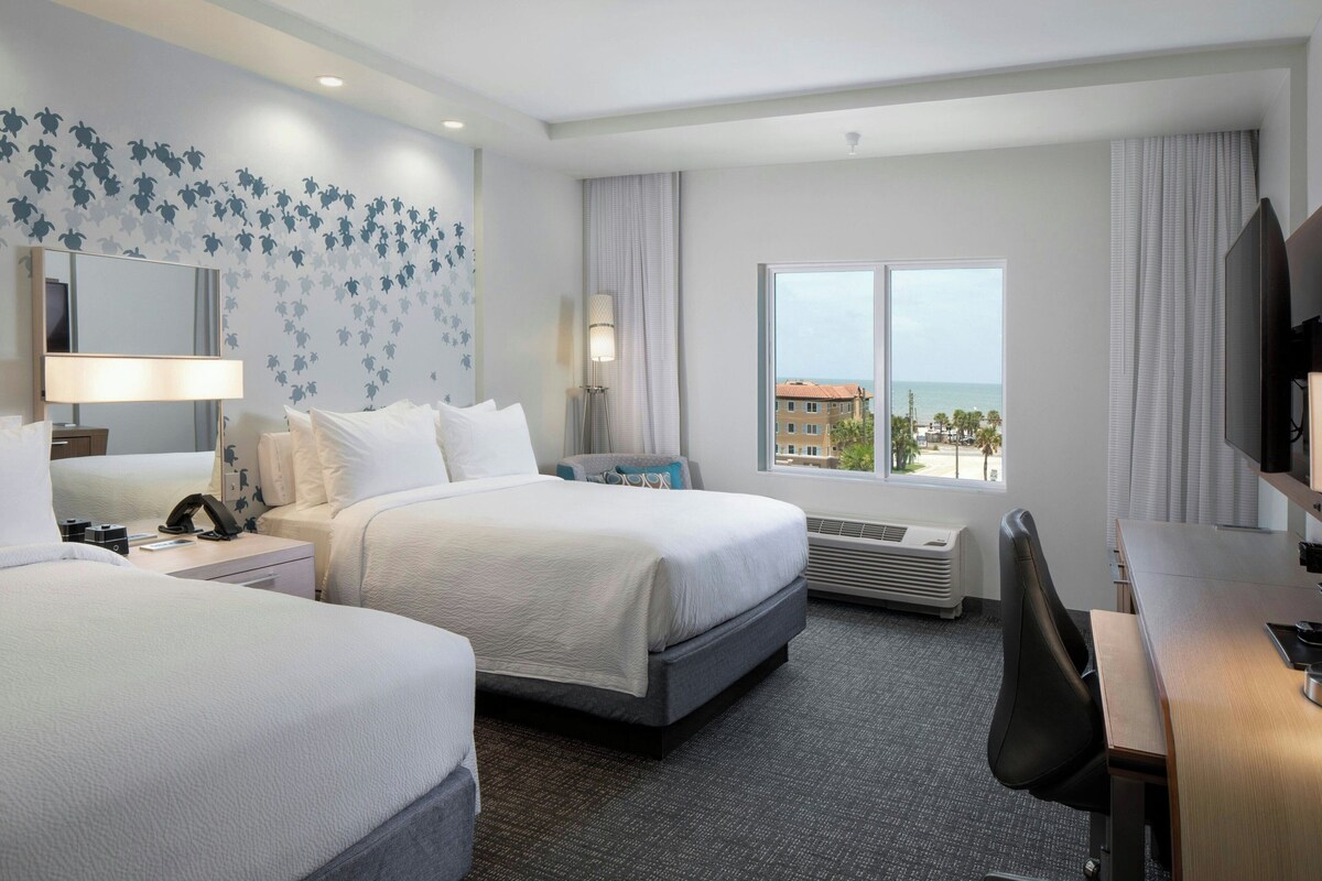 Une chambre d’hôtel avec deux lits, un bureau, une télévision, une grande fenêtre donnant sur la mer et un mur orné d’un motif floral.