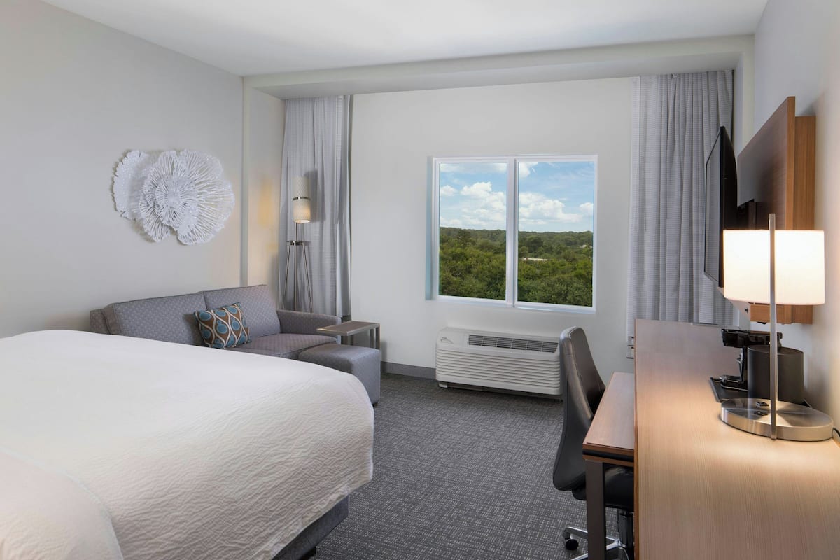 Une chambre d’hôtel avec un grand lit, un bureau, une chaise, une télévision, une lampe et une fenêtre donnant sur un paysage verdoyant.