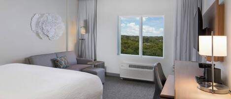 Habitación de hotel con una cama grande, un escritorio, una silla, un televisor, una lámpara y una ventana con vistas a zonas verdes.