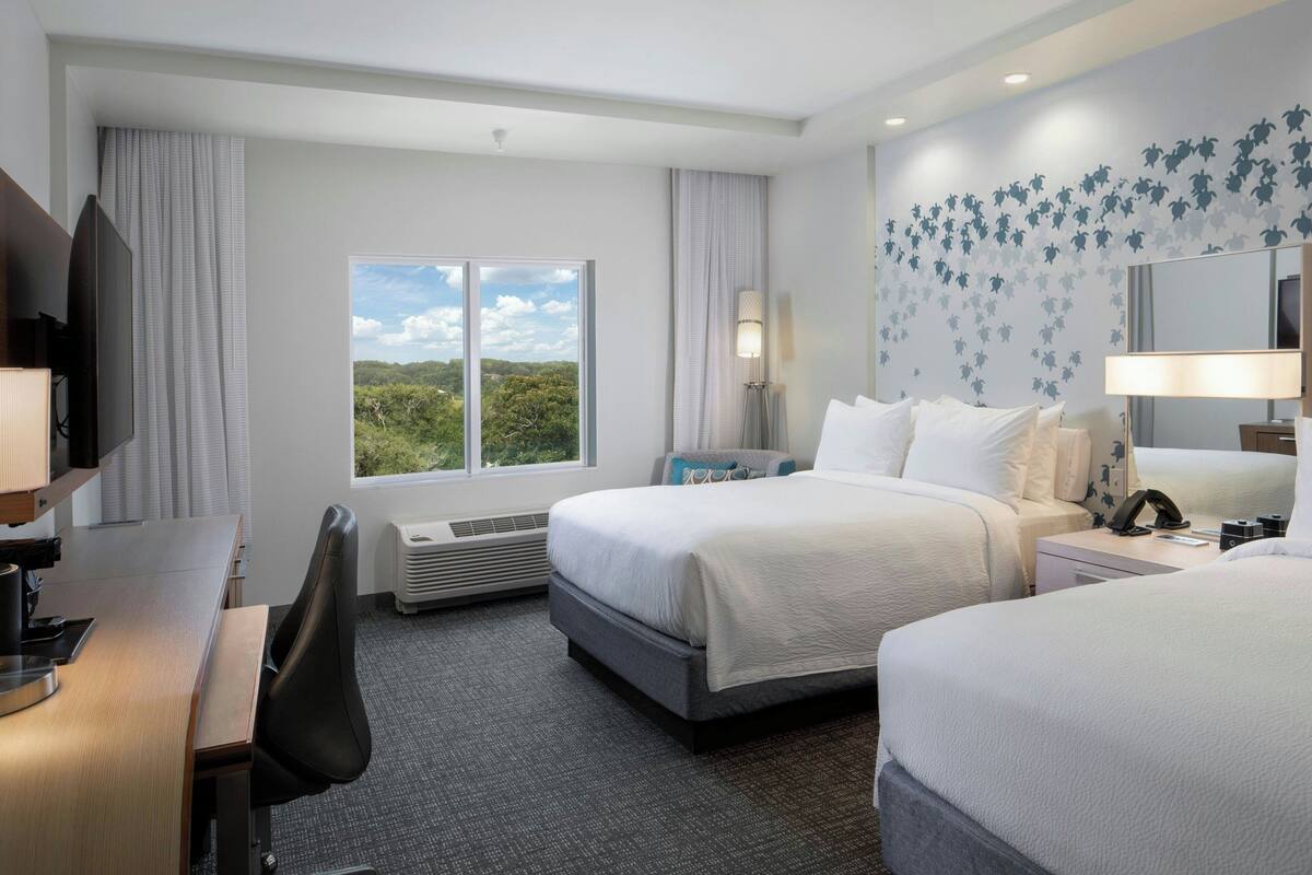 Une chambre d’hôtel avec deux lits, un bureau, une chaise, une télévision, une lampe et une fenêtre donnant sur un paysage verdoyant.