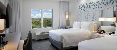 A hotel room with two beds, a desk, a chair, a TV, a lamp, and a window with a view of greenery.