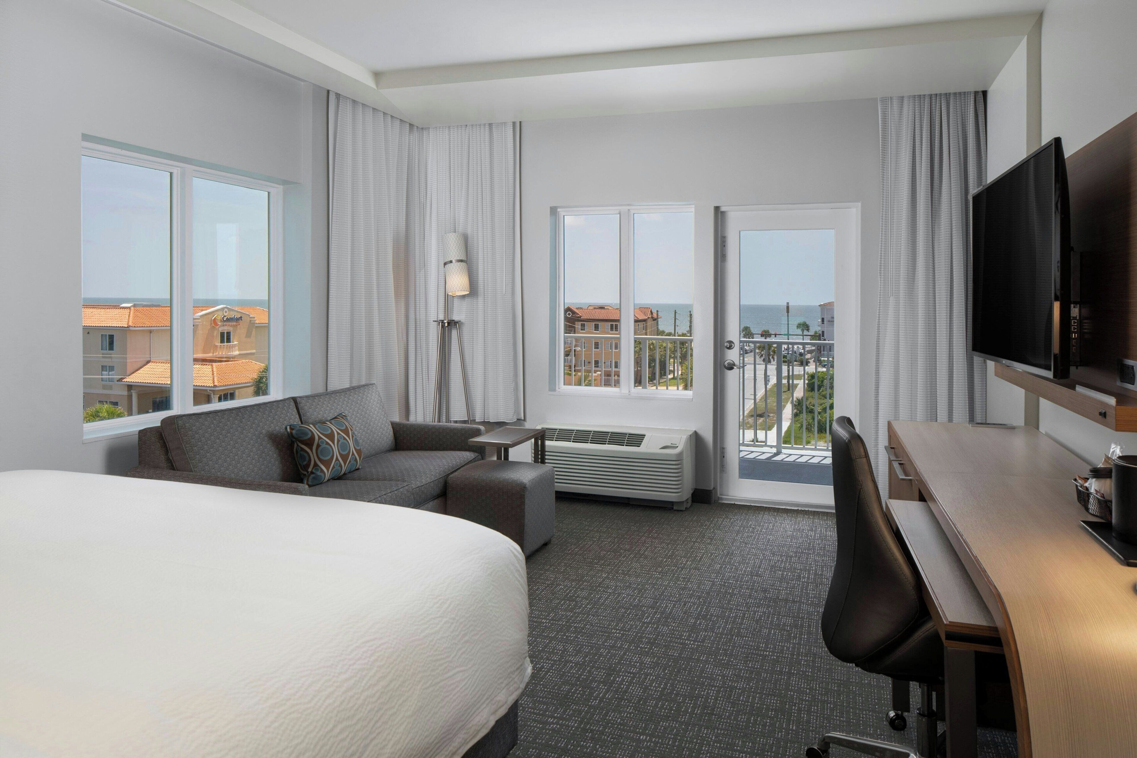 A hotel room with a large bed, a desk, a chair, a sofa, and a TV. There are two windows and a door leading to a balcony.