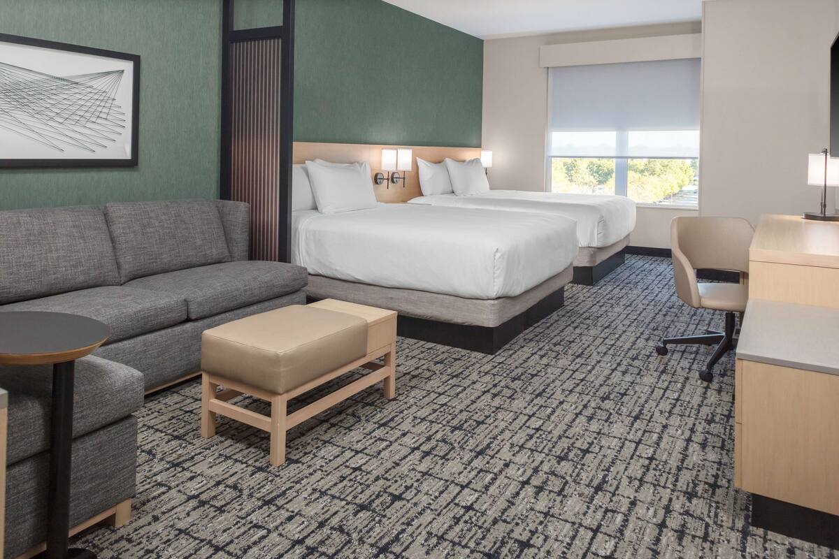 Habitación de hotel con un sofá gris, una mesita redonda pequeña, un banco de madera, dos camas individuales, un escritorio con una silla y un ventanal grande.