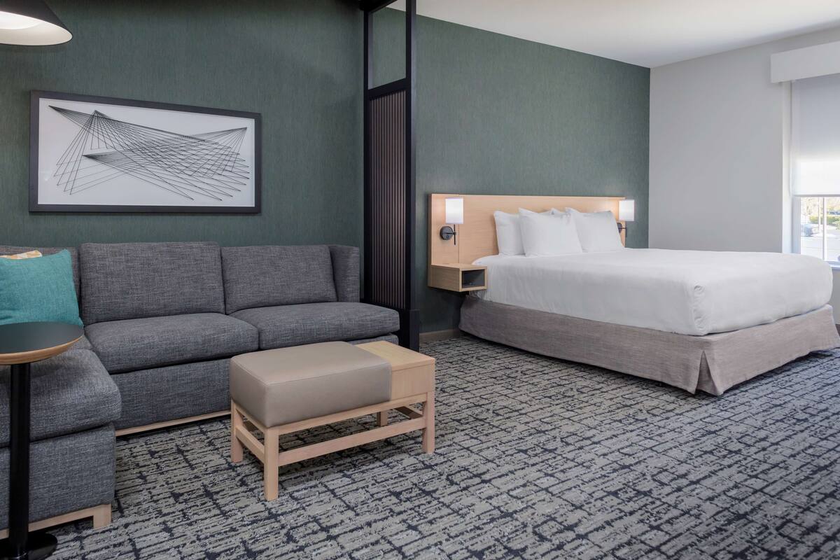 Una habitación de hotel moderna con un sofá gris, un banco de madera, una cama grande, una obra de arte abstracta enmarcada y una ventana con vistas.