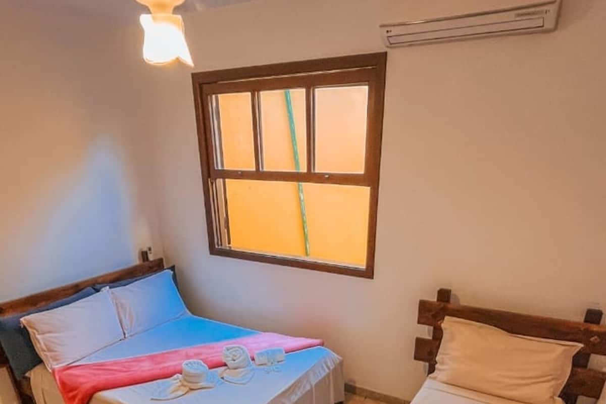 A small, cozy room with two single beds, a ceiling fan, a window with blinds, and a wall-mounted air conditioner.