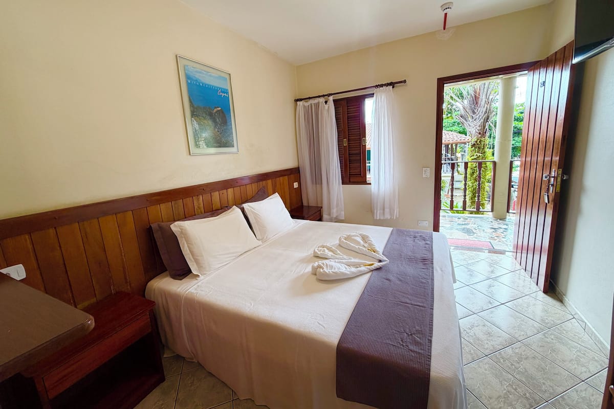 A hotel room with a bed, a wooden headboard, a bedside table, a window with curtains, and a door leading to an outdoor area.