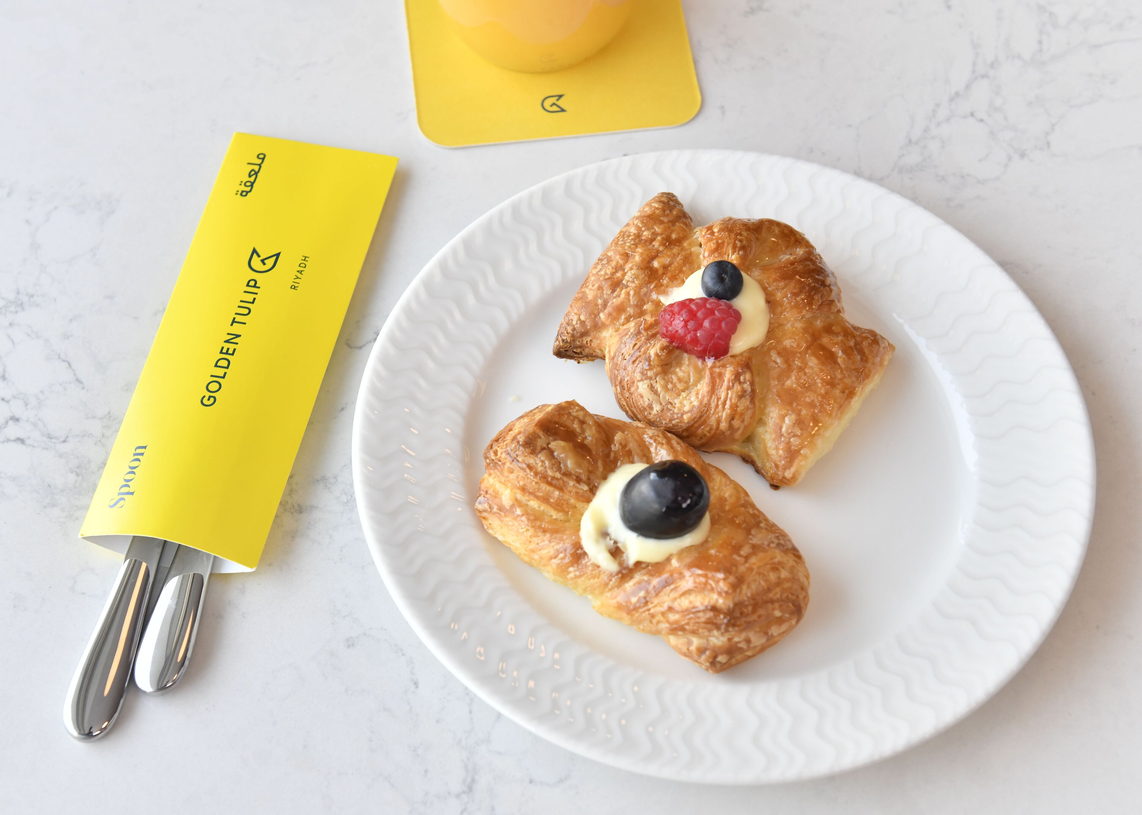 A white plate with two pastries, one with a blueberry and the other with a raspberry, and a yellow spoon on a white marble surface.