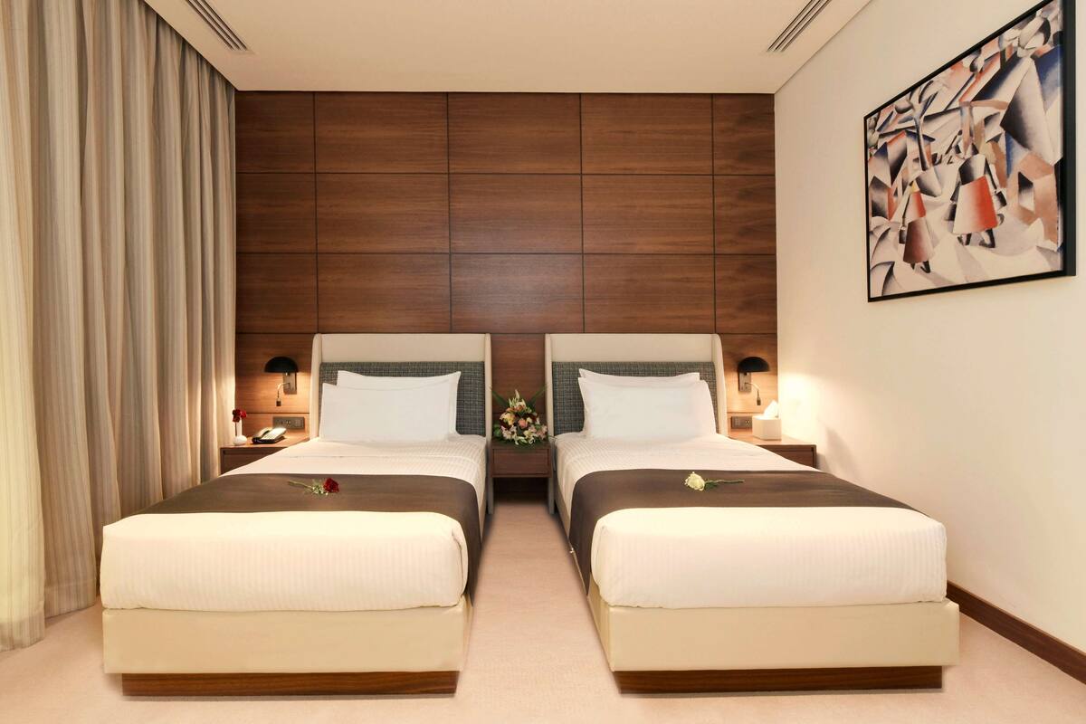 A hotel room with two beds, wooden headboards, a large abstract painting, and a nightstand with a lamp.