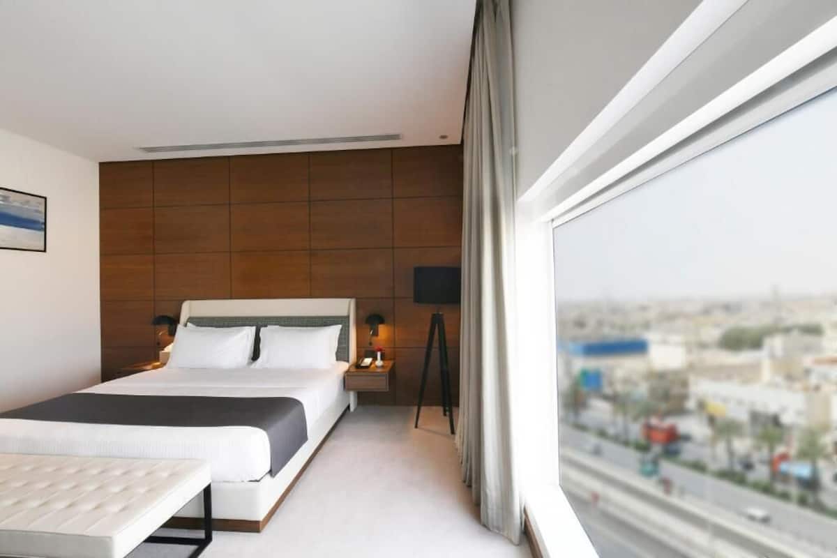 A modern hotel room with a large bed, a bedside table, a lamp, and a view of the city through a large window.