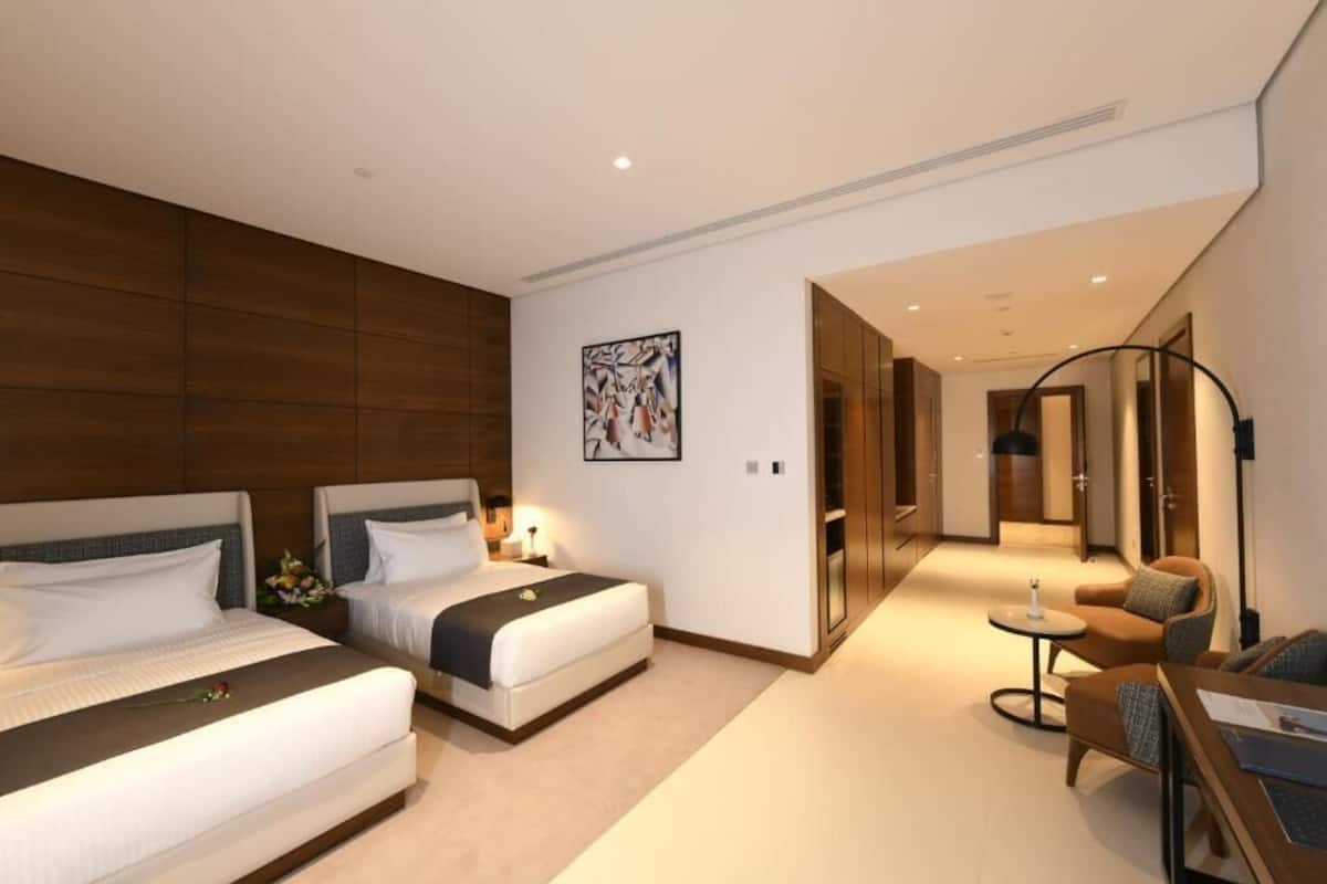 A hotel room with two beds, a sitting area with a sofa and a small table, and a hallway leading to another room.