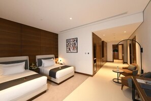 Une chambre d’hôtel avec deux lits, un coin salon avec un canapé et une petite table, et un couloir menant à une autre pièce.