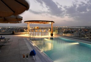Un espace piscine aménagé sur le toit, avec un bar et des chaises longues, offrant une vue sur le paysage urbain au crépuscule.