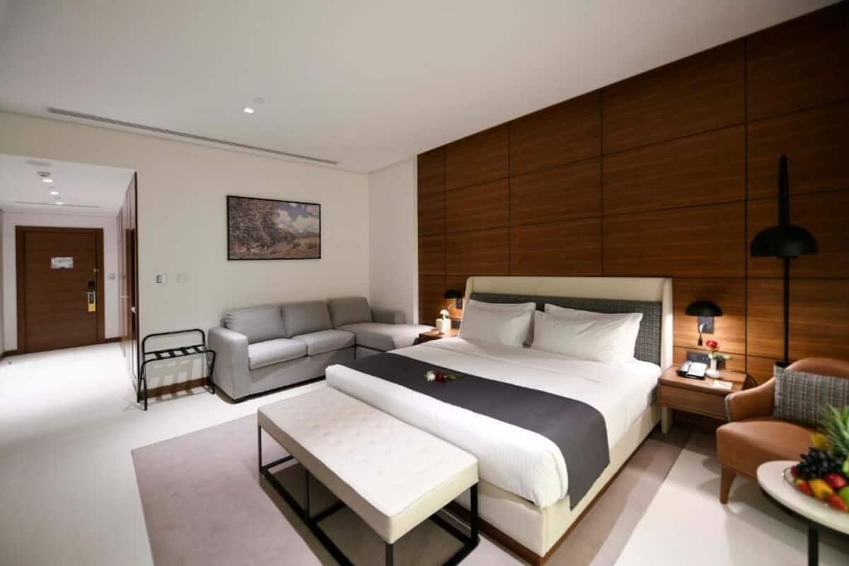 A modern hotel room with a large bed, a sofa, a bench, and a small table with fruit.