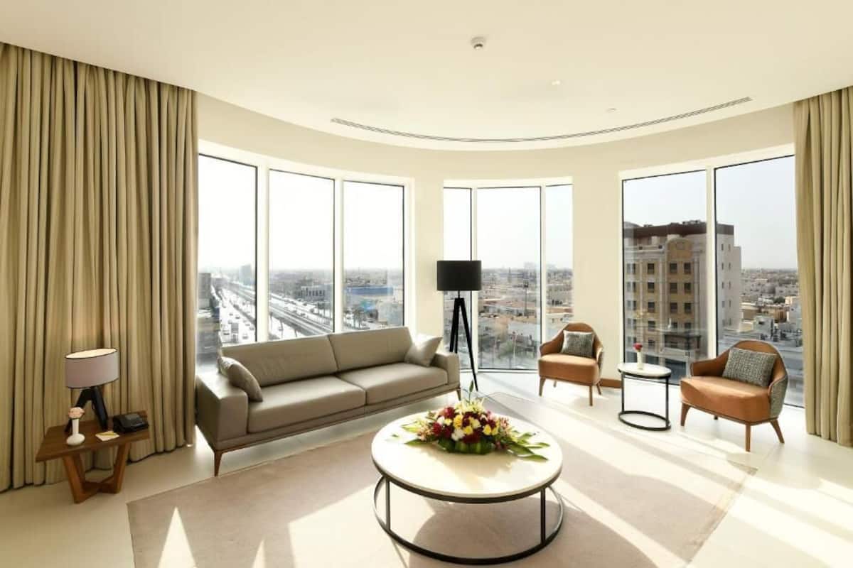 A modern, well-lit living room with a large sofa, a coffee table with flowers, and a view of the city through large windows.