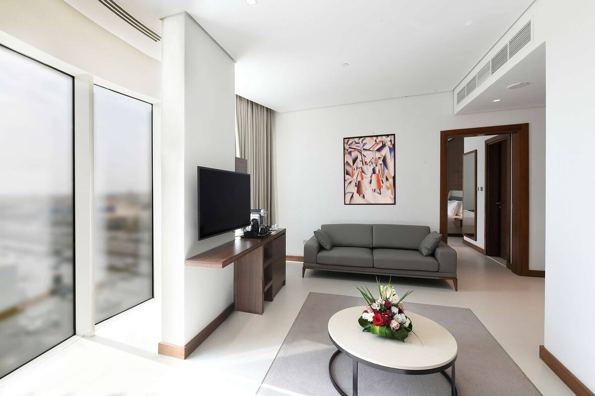 A modern hotel room with a large window, a flat-screen TV, a gray sofa, a coffee table with a floral arrangement, and a painting on the wall.
