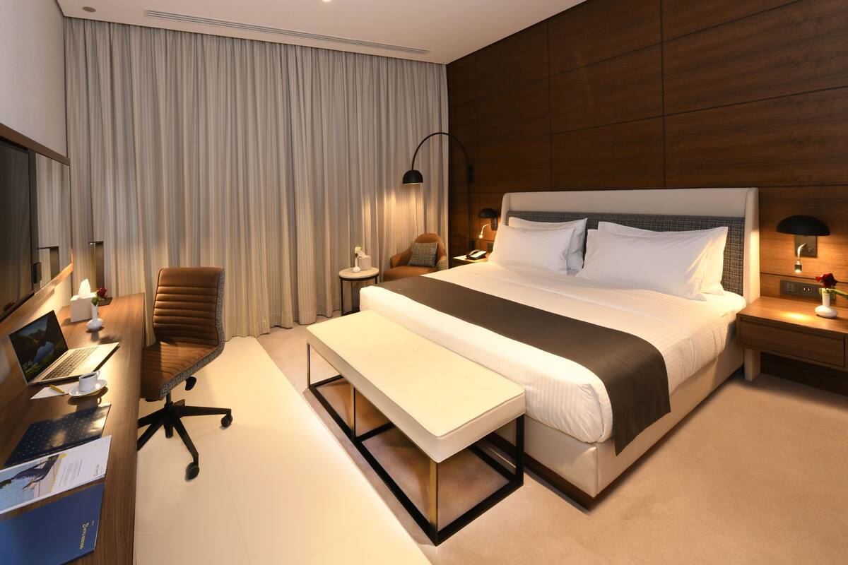 A modern hotel room with a large bed, a desk with a laptop, a chair, and a bedside table with a lamp.