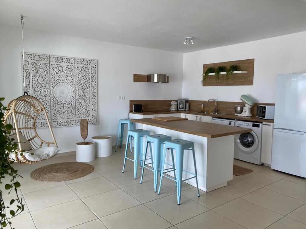 Una cocina moderna con una isla central, taburetes de bar, lavadora y una silla colgante.