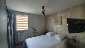 Een slaapkamer met een bed, een raam met zonwering, een radiator, een plafondventilator, een nachtkastje met een lamp en een aan de muur hangend kunstwerk.