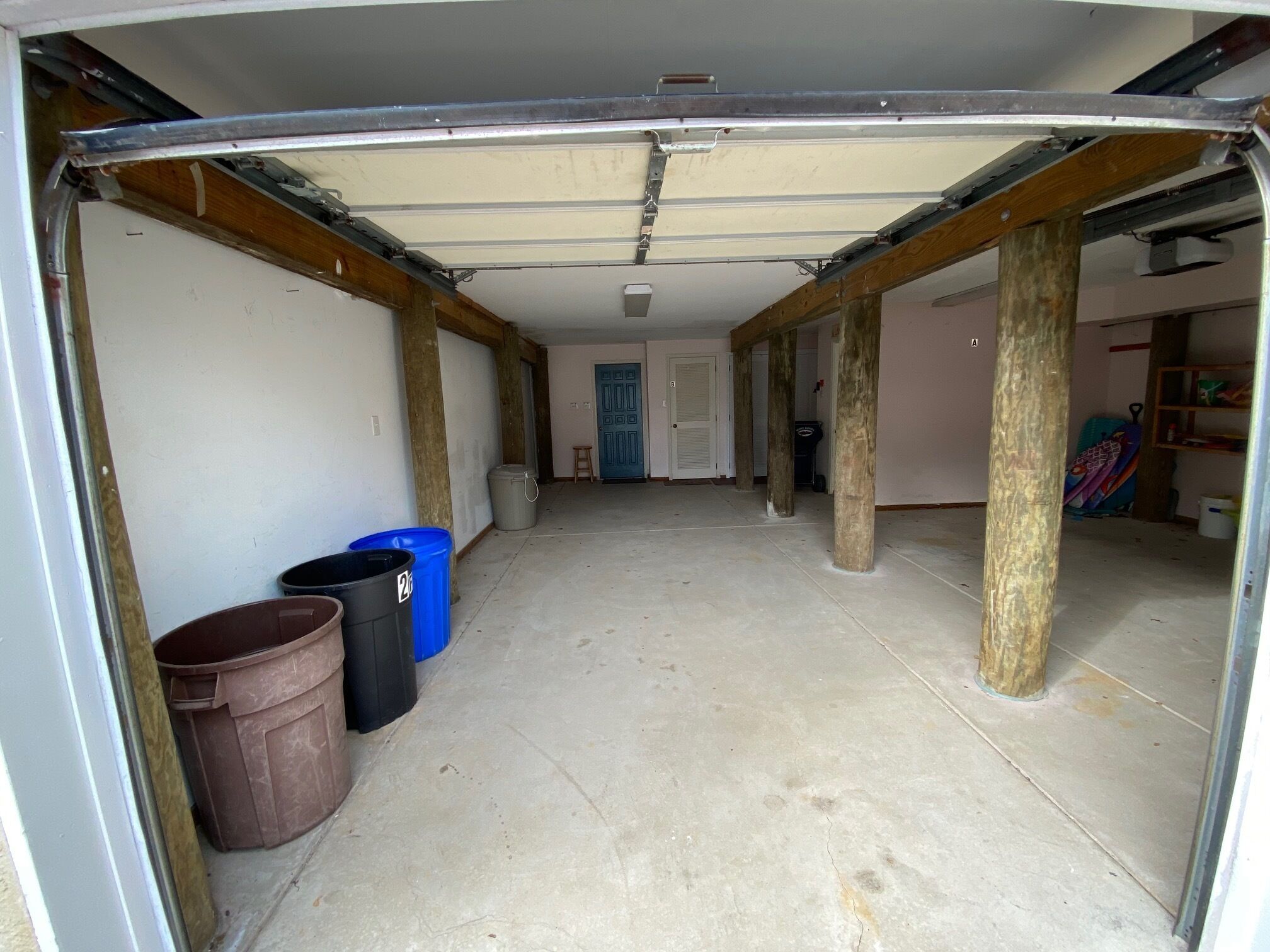 A garage with concrete floor, wooden beams, and a blue trash bin.
