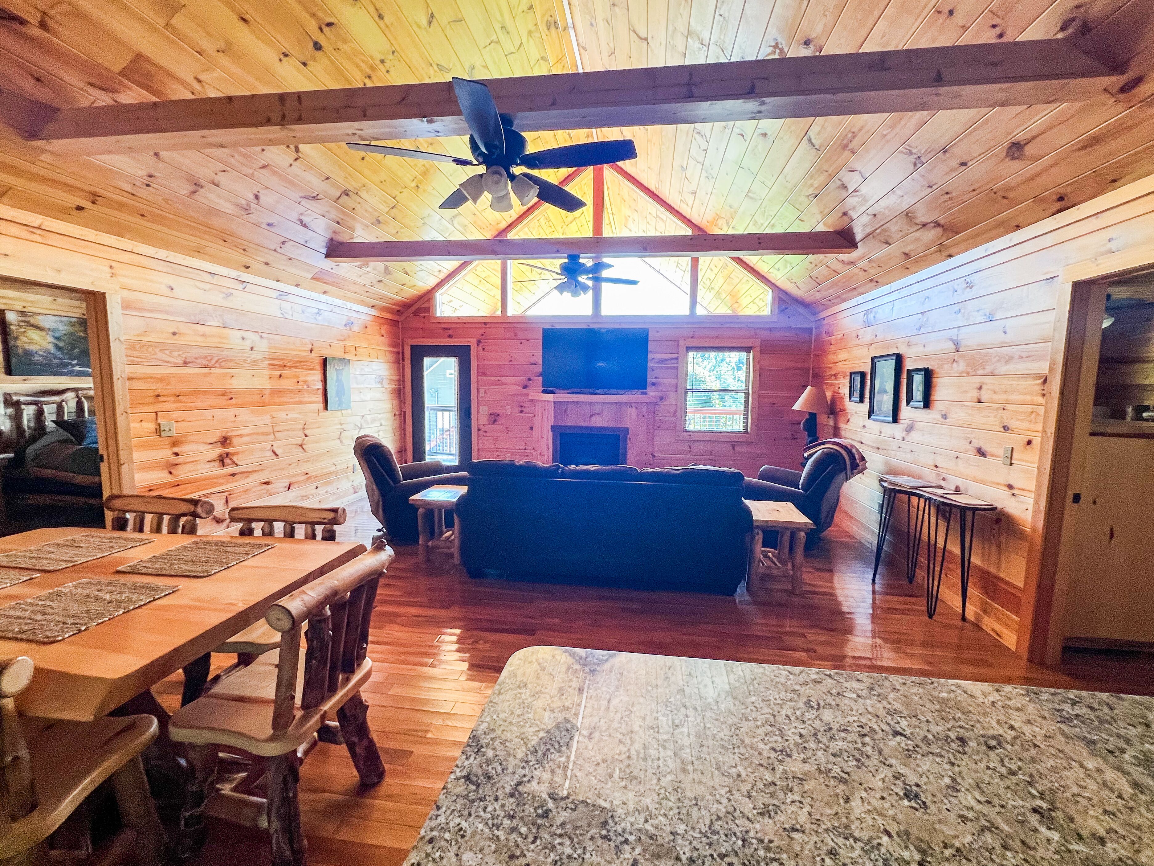 L’intérieur chaleureux d’un chalet avec un plancher en bois, une cheminée, un ventilateur de plafond et un coin repas doté de meubles en bois.