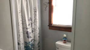 A bathroom with a toilet, a shower curtain with floral patterns, a window with a wooden frame, and a bottle of hand soap on the toilet tank. - Sunny Cottage at Cozy Nook (Lake George)