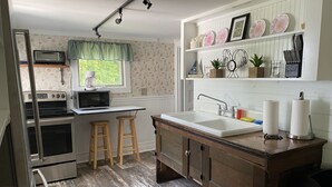 A compact kitchen with a microwave, stove, and wooden stools. It features a window with a green valance, a sink with paper towels, and decorative plates on the wall. - Hens & Chickens Cottage at Cozy Nook (Lake George)
