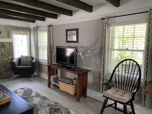 Una sala de estar con un televisor sobre un mueble de madera, una silla negra con cojín, una pared de piedra y una ventana con cortinas estampadas.